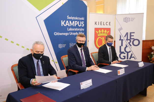 Trzech mężczyzn w garniturach i maskach siedzi przy stole. Na stole dokumenty i długopisy. Tło to banery z logo instytucji.