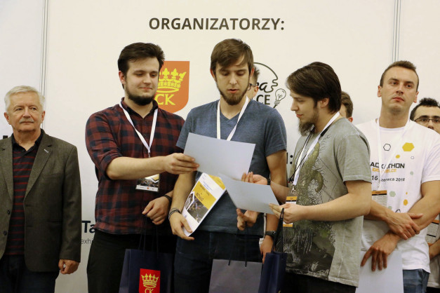 Mężczyźni w koszulkach i marynarkach stoją przed napisem "ORGANIZATORZY". Trzymają dokumenty i torby.