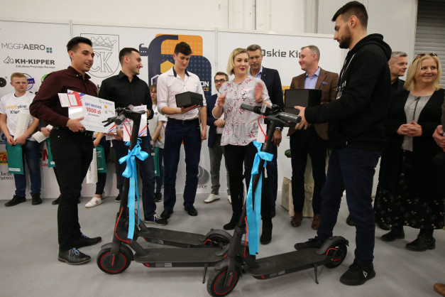 Grupa ludzi odbiera nagrody, w tym hulajnogi elektryczne i bony podarunkowe. Kontekst: wydarzenie lub konkurs.