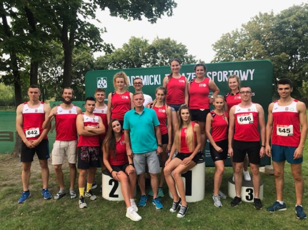 Grupa sportowców w strojach sportowych pozuje do zdjęcia na tle zielonego banera. W tle widać drzewa.