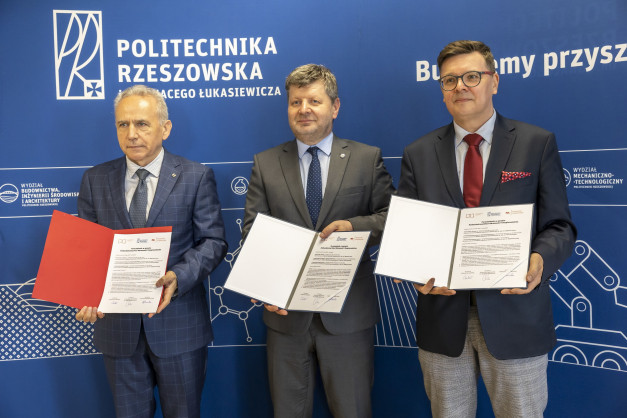 Trzech mężczyzn w garniturach pozuje z dokumentami przed niebieskim tłem z logo Politechniki Rzeszowskiej.