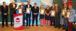 Grupa osób w garniturach odbiera nagrody. Kluczowe obiekty: mężczyźni, kobiety, dyplomy, statuetki, podium.