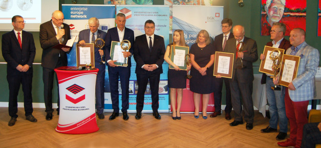 Grupa osób w garniturach odbiera nagrody. Kluczowe obiekty: mężczyźni, kobiety, dyplomy, statuetki, podium.