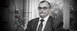 Mężczyzna w okularach i garniturze stoi obok okna. W tle widoczne są rury i drzewa.