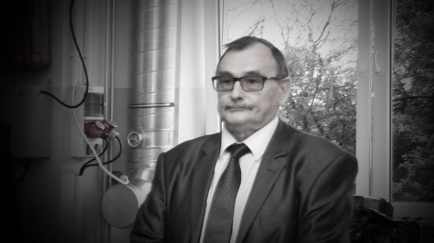 Mężczyzna w okularach i garniturze stoi obok okna. W tle widoczne są rury i drzewa.