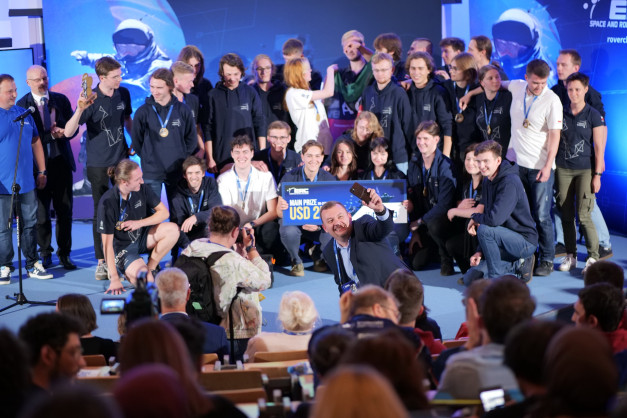 Grupa młodych ludzi w strojach z logo, odbiera nagrodę. W tle ekran z astronautą.