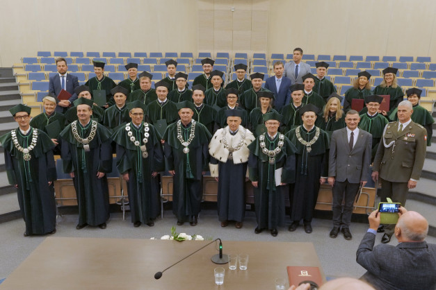 Grupa osób w togach akademickich i biretach pozuje do zdjęcia w sali wykładowej.