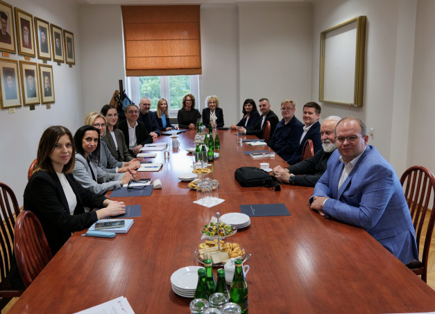 Grupa osób siedzi przy stole konferencyjnym. Na stole są dokumenty, napoje i przekąski.