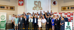 Grupa ludzi pozuje do zdjęcia na tle banerów z napisem "50 1969 2019". Kluczowe obiekty: ludzie, banery, godło Polski.