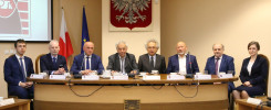 Osiem osób siedzi przy stole konferencyjnym. Na ścianie wisi godło Polski.