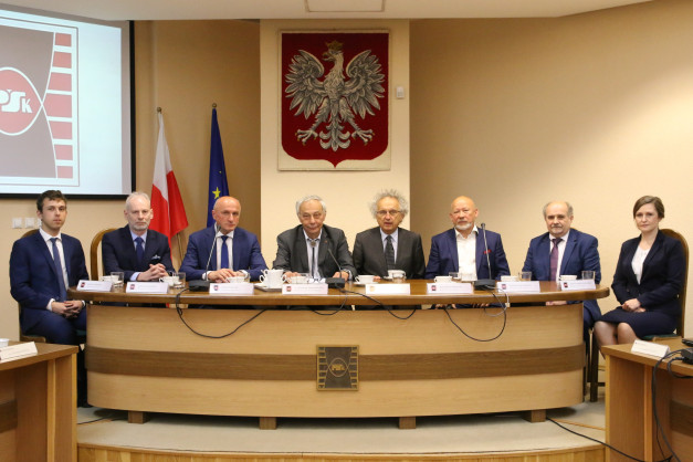 Osiem osób siedzi przy stole konferencyjnym. Na ścianie wisi godło Polski.