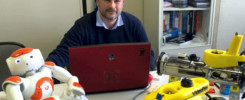 Mężczyzna z robotami i laptopem. Na stole robot humanoidalny, żółty pojazd podwodny i czerwony laptop.