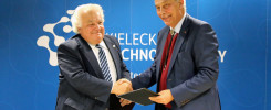 Dwóch mężczyzn w garniturach ściska sobie dłonie na tle niebieskiej ściany z logo Kieleckiego Parku Technologicznego.