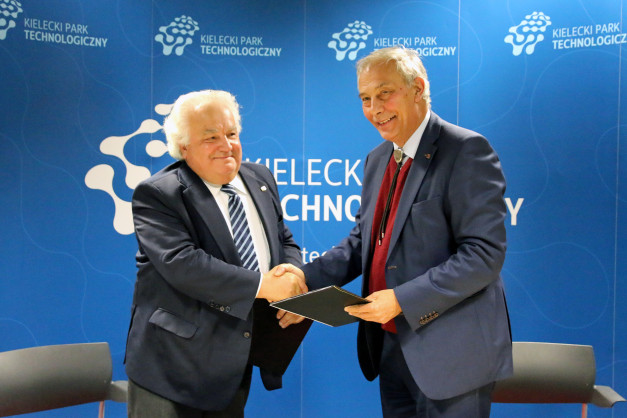 Dwóch mężczyzn w garniturach ściska sobie dłonie na tle niebieskiej ściany z logo Kieleckiego Parku Technologicznego.