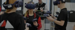 Osoby w goglach VR i kontrolerach w rękach, w pomieszczeniu z wyposażeniem. Kontekst: technologia, edukacja.