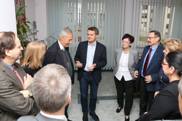 Grupa osób rozmawia w pomieszczeniu. Widać mężczyzn w garniturach i kobiety w eleganckich strojach.