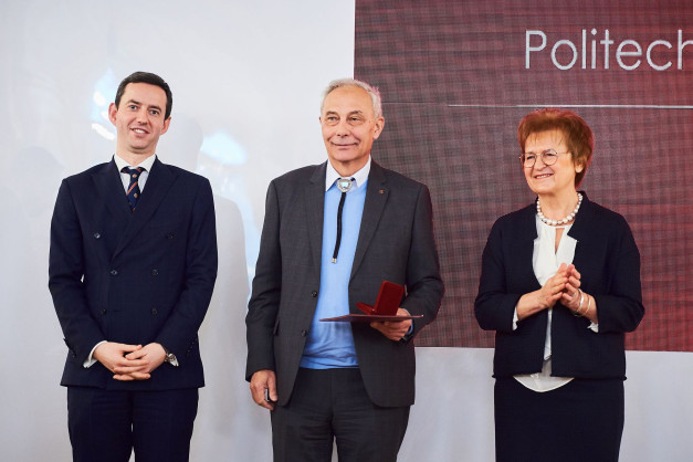 Trzy osoby stoją na scenie przed napisem "Politechnika". Mężczyzna w garniturze trzyma czerwone pudełko.