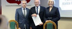 Trzy osoby w eleganckich strojach stoją przed herbem Polski. Mężczyzna w środku trzyma dokument.