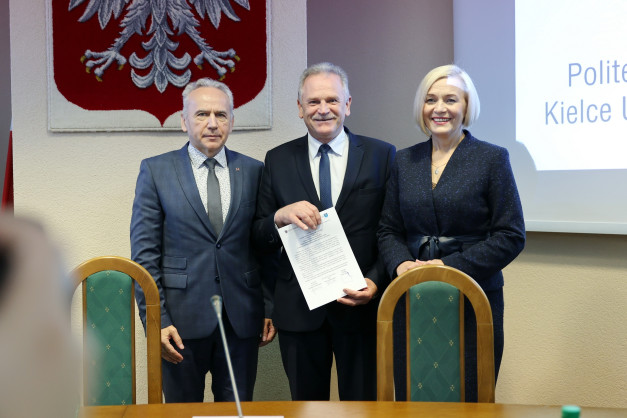 Trzy osoby w eleganckich strojach stoją przed herbem Polski. Mężczyzna w środku trzyma dokument.