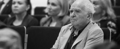 Starszy mężczyzna w marynarce siedzi w sali. W tle widać inne osoby.