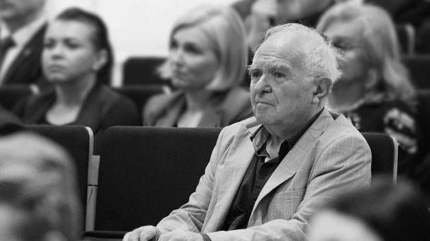 Starszy mężczyzna w marynarce siedzi w sali. W tle widać inne osoby.