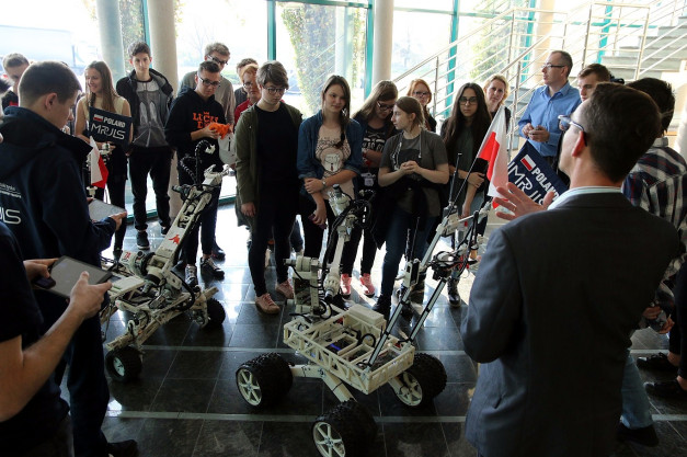 Grupa młodych ludzi ogląda roboty. W tle widać flagę Polski.