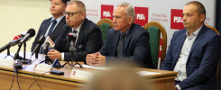 Czterech mężczyzn w garniturach siedzi przy stole konferencyjnym z mikrofonami.