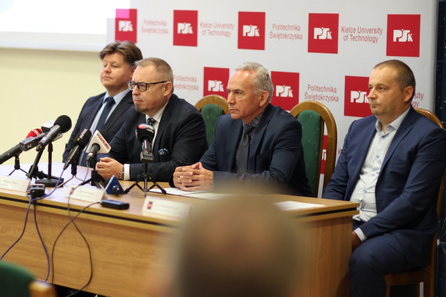 Czterech mężczyzn w garniturach siedzi przy stole konferencyjnym z mikrofonami.