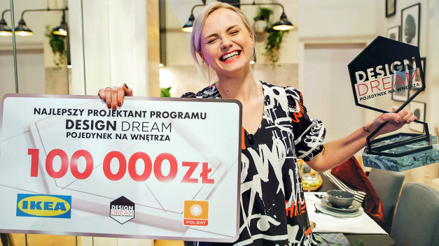 Kobieta trzyma tablicę z napisem "Najlepszy projektant programu DESIGN DREAM 100 000 zł" i statuetkę.