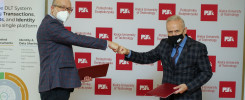 Dwóch mężczyzn w garniturach i maseczkach wymienia uścisk pięści na tle ścianki z logo Politechniki Świętokrzyskiej.