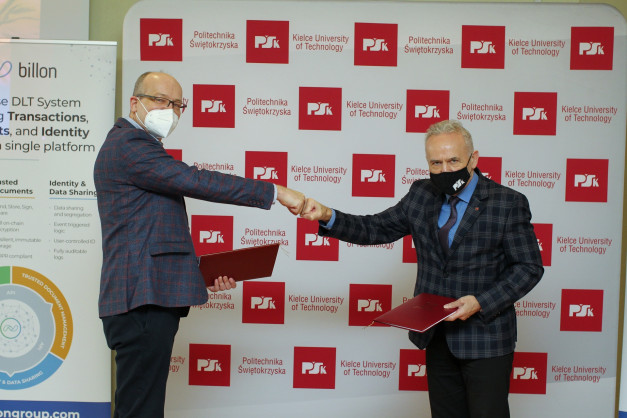 Dwóch mężczyzn w garniturach i maseczkach wymienia uścisk pięści na tle ścianki z logo Politechniki Świętokrzyskiej.