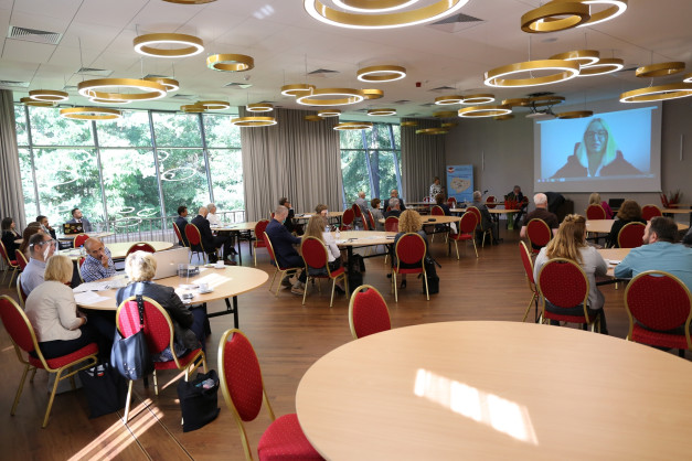 Sala konferencyjna z ludźmi siedzącymi przy stołach. Kluczowe obiekty: stoły, krzesła, ekran projekcyjny, okna.