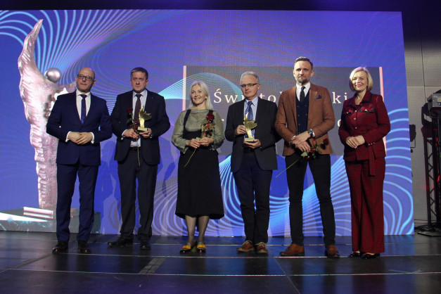 Sześć osób na scenie, trzymających statuetki i róże. Tło to niebieski ekran z falistym wzorem.