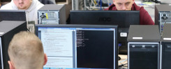 Osoby pracujące przy komputerach w sali lekcyjnej. Kluczowe obiekty: monitory, komputery, kable. Kontekst: nauka, technologia.