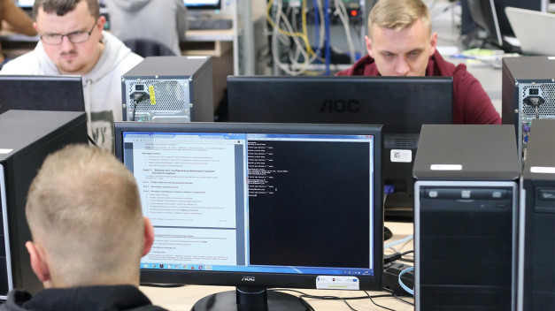 Osoby pracujące przy komputerach w sali lekcyjnej. Kluczowe obiekty: monitory, komputery, kable. Kontekst: nauka, technologia.