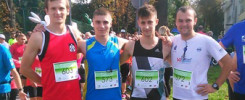 Czterech młodych mężczyzn w strojach sportowych pozuje do zdjęcia podczas biegu. W tle widać innych uczestników i drzewa.