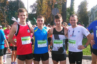 Czterech młodych mężczyzn w strojach sportowych pozuje do zdjęcia podczas biegu. W tle widać innych uczestników i drzewa.