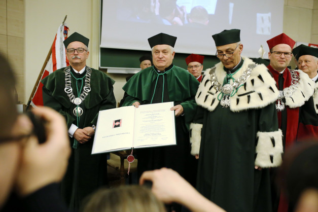 Uroczystość akademickia. Kluczowe obiekty: dyplom, togą, łańcuchy. Kontekst: ceremonia akademicka.