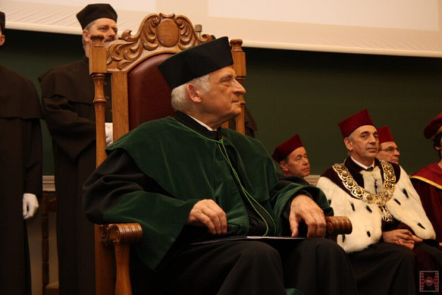 Mężczyzna w togach akademickich siedzi na krześle. W tle inni mężczyźni w togach.