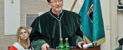 Mężczyzna w todze akademickiej przemawia przy mównicy. Obok stoi kobieta w szarfie. Kontekst: uroczystość akademicka.
