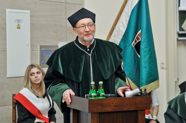Mężczyzna w todze akademickiej przemawia przy mównicy. Obok stoi kobieta w szarfie. Kontekst: uroczystość akademicka.