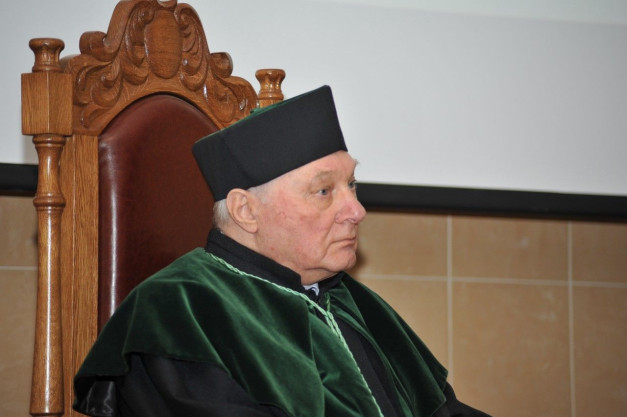 Starszy mężczyzna w togach akademickich siedzi na ozdobnym krześle.