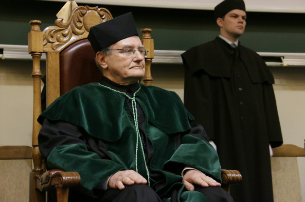 Mężczyzna w togach akademickich siedzi na ozdobnym krześle. Za nim stoi drugi mężczyzna w czarnej todze.