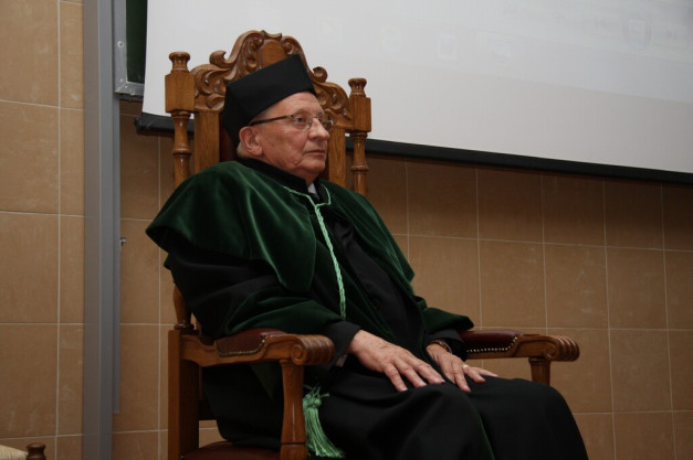 Starszy mężczyzna w todze i birecie siedzi na ozdobnym drewnianym krześle. Kontekst: uroczystość akademicka.