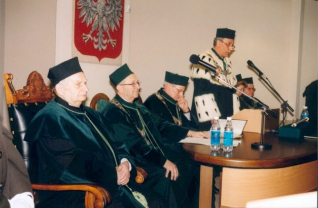 Uroczystość akademicka. W sali siedzą profesorowie w togach, jeden przemawia. Kontekst: ceremonia.