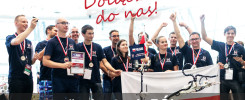 Grupa młodych ludzi w koszulkach z logo, z medalami i dyplomem, trzymająca flagę Polski.