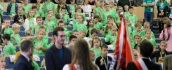 Grupa dzieci w zielonych koszulkach siedzi na widowni. W tle dorośli.