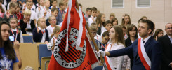 Młodzi ludzie w mundurkach szkolnych i dorośli w sali. Na pierwszym planie sztandar z godłem.