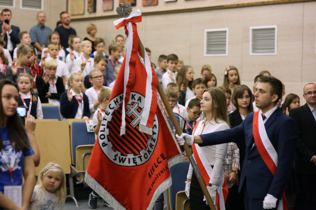 Młodzi ludzie w mundurkach szkolnych i dorośli w sali. Na pierwszym planie sztandar z godłem.