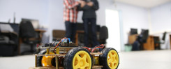 Zbudowany robot mobilny z żółtymi kołami i dwoma chłopcami w tle. Kontekst: warsztaty lub pracownia.